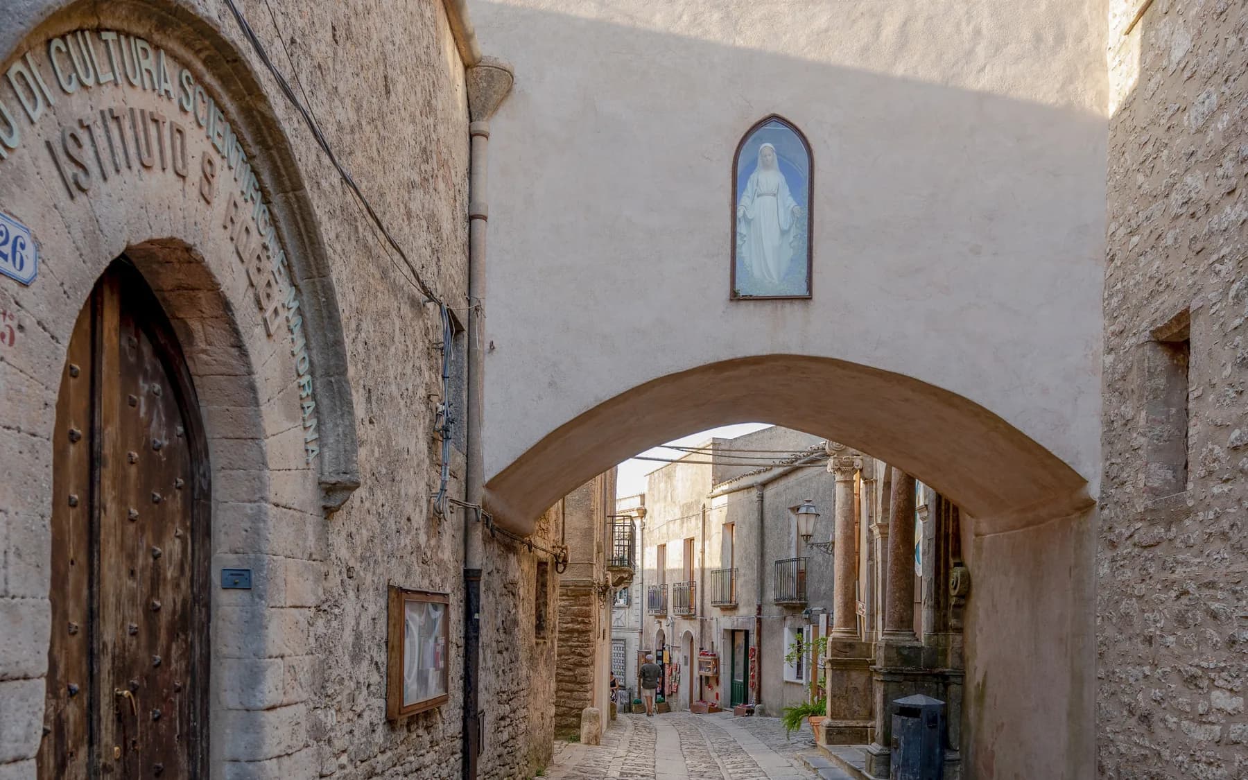 A stone archway over a cobbled lane in Erice, with a studded wooden door to one side, a small painted shrine above the arch and a figure walking towards the shops beyond