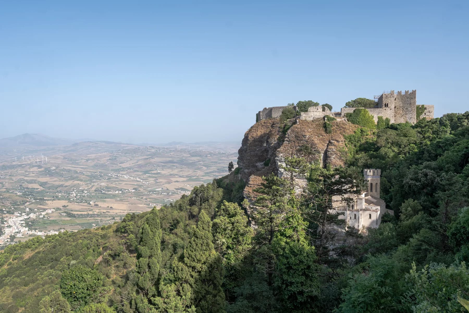 The castle of Erice on a wooded clifftop, with a small turreted building below it and farmland stretching to the horizon