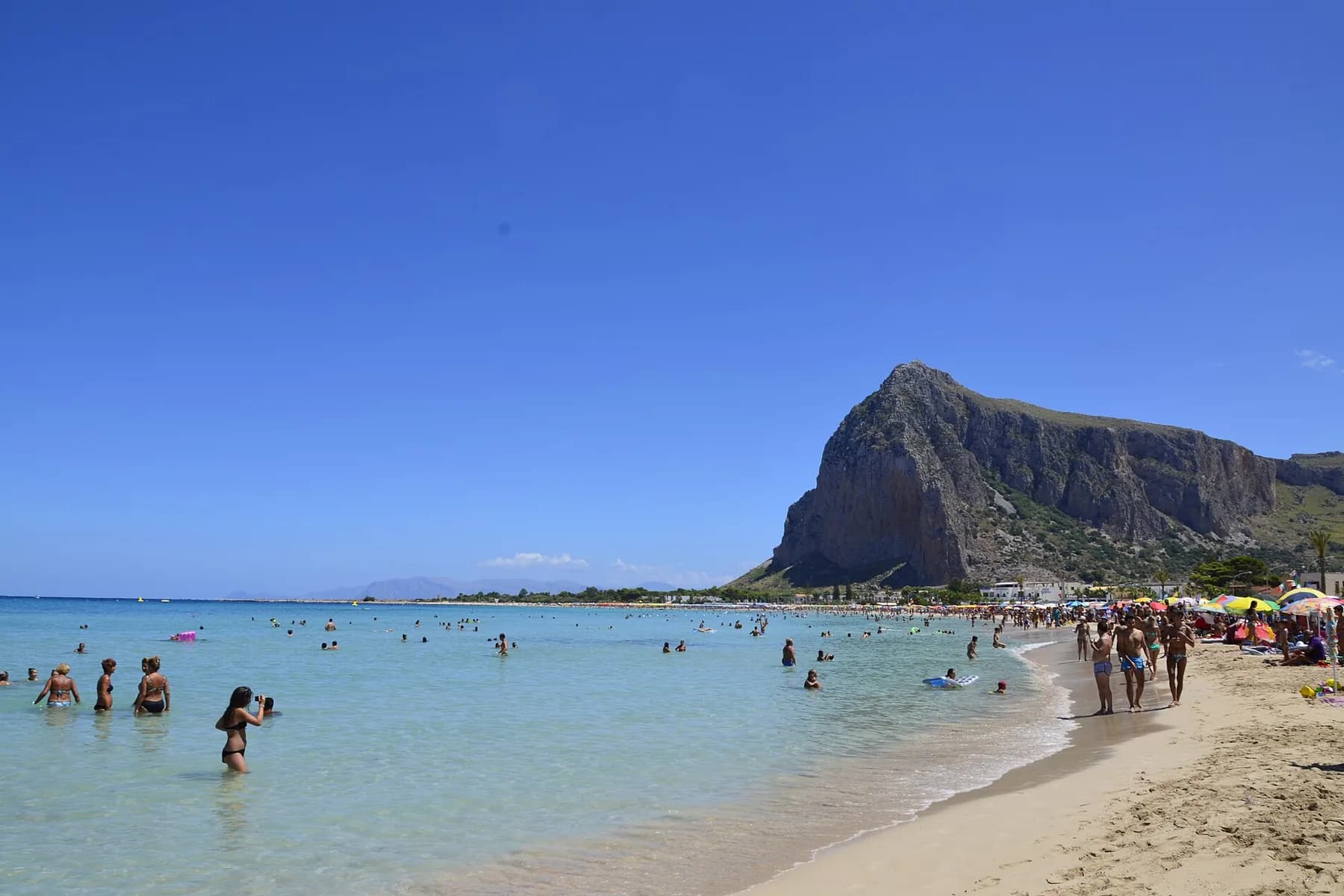 Shallow turquoise water with bathers along a curve of pale sand, and a high rocky headland rising at the far end of the bay
