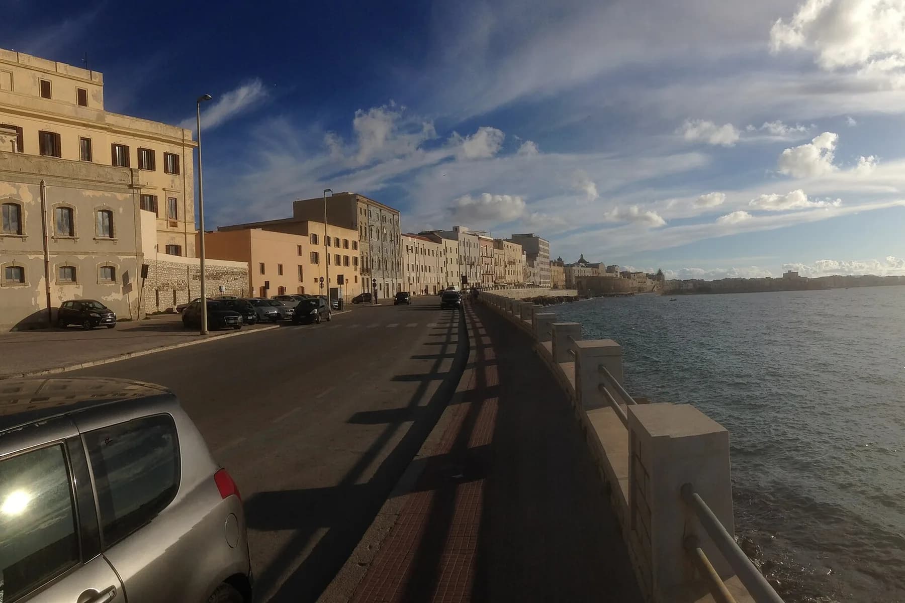The seafront road running along the old town of Trapani, with the sea wall on one side and the open sea beyond under a blue sky