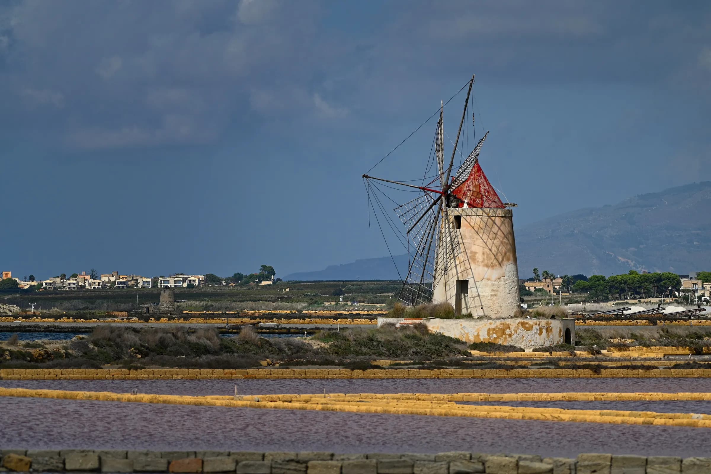 A windmill with a red cap standing among the Trapani salt pans, pink and ochre basins in front of it and mountains under a dark sky