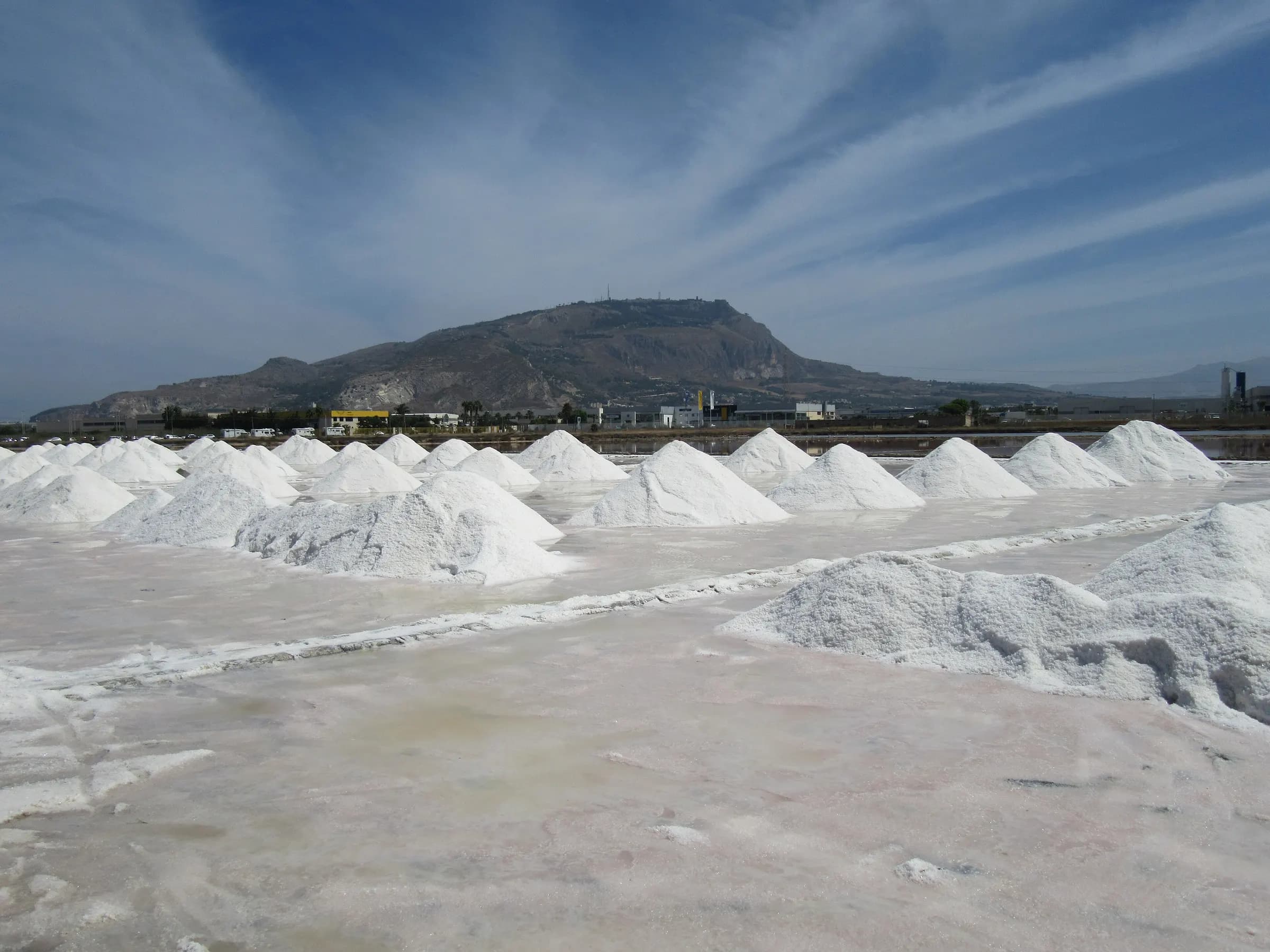 Heaps of white salt along the banks of the Trapani salt pans, with Mount Erice rising behind under a streaked blue sky