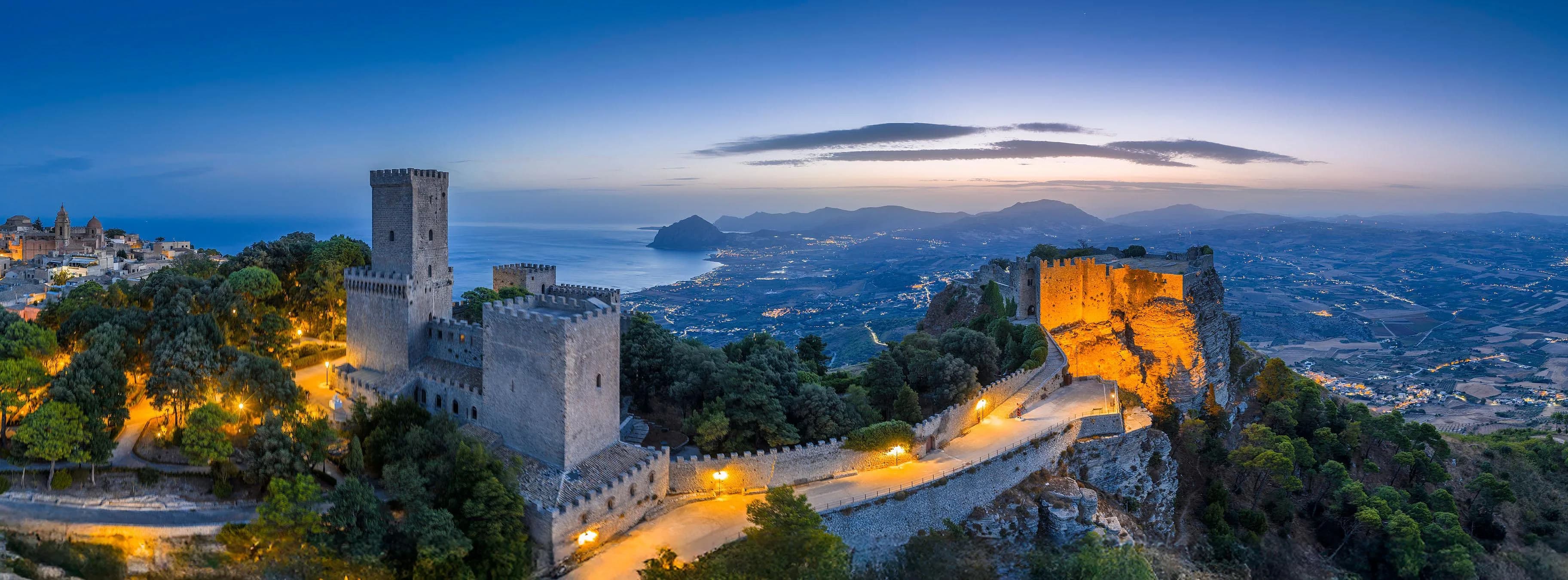 The walls and castle of Erice lit at dusk, with lamps along the path below them and the gulf and coastline stretching away beneath