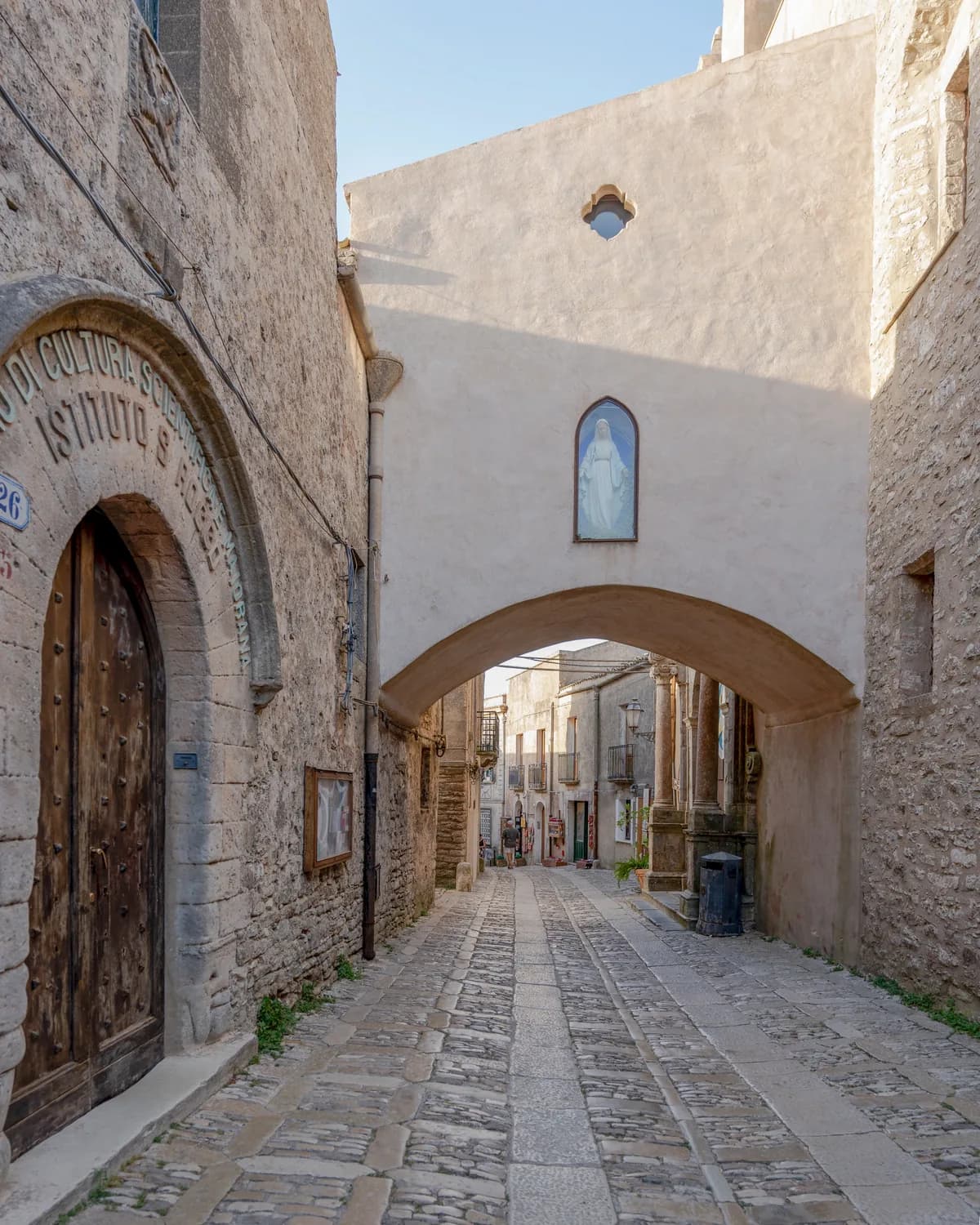 A cobbled lane running under a stone arch, with a studded wooden door on the left and a small painted shrine set into the arch above