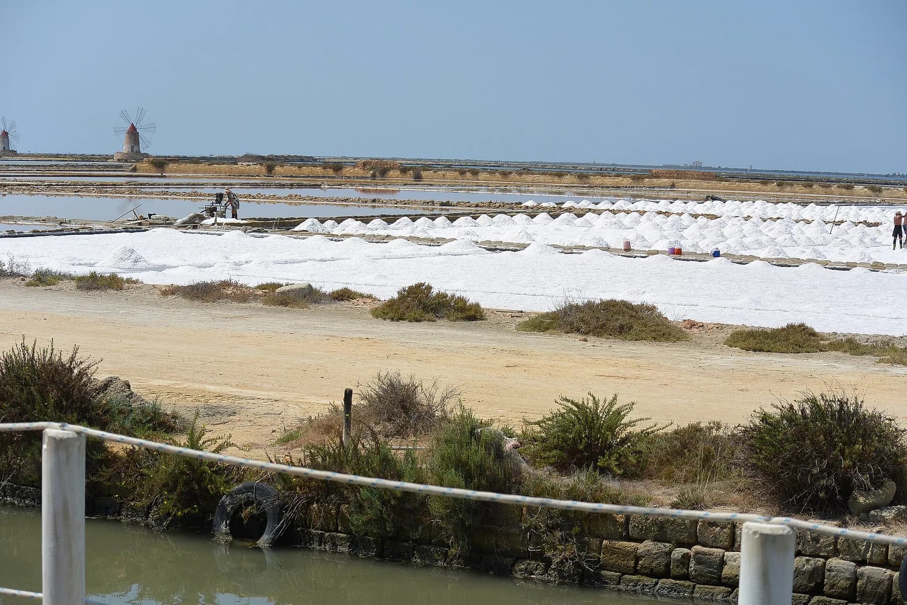 Rows of white salt heaped between the basins on a bright day, with workers out on the banks and a windmill on the far horizon