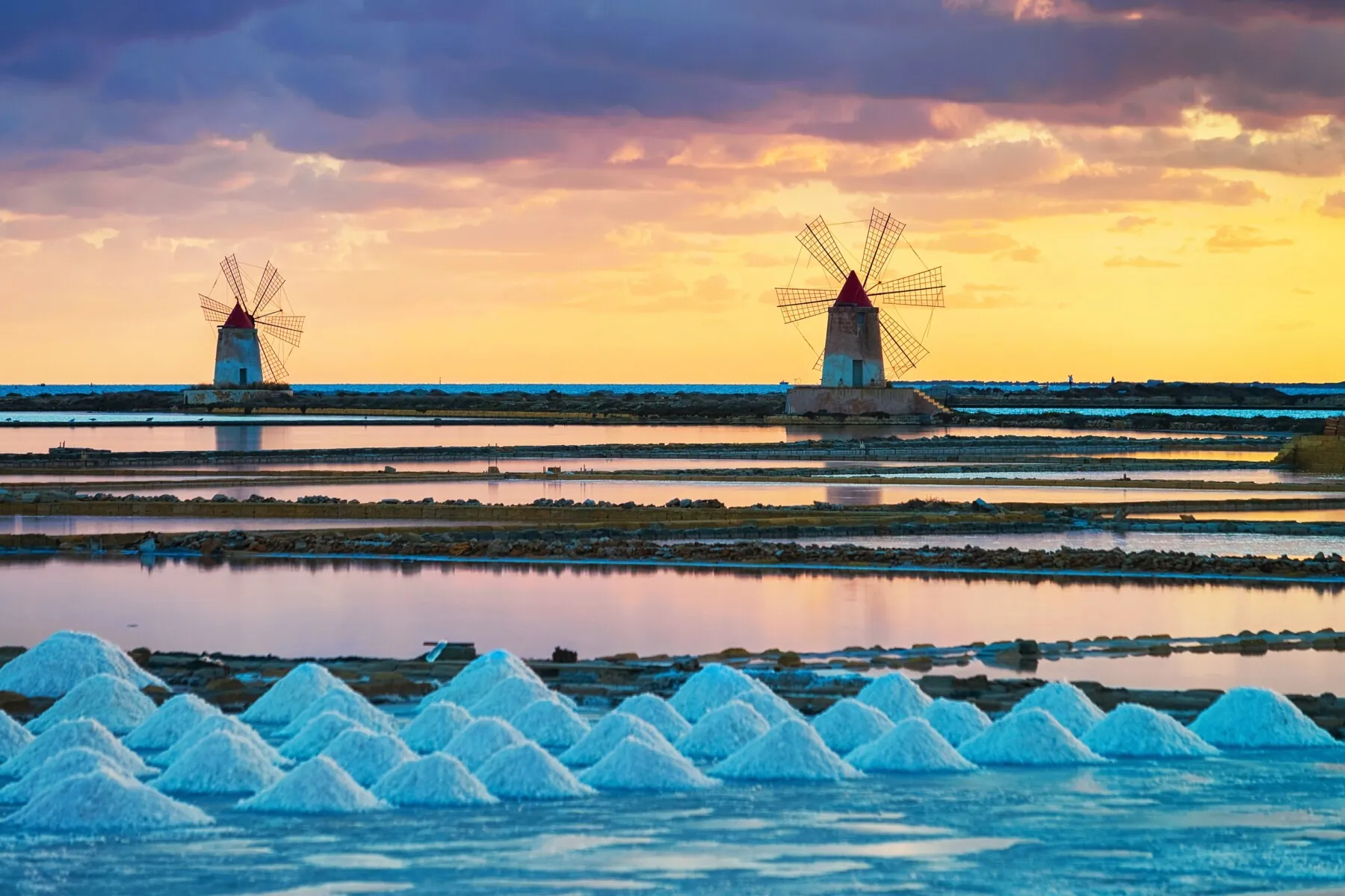 Two windmills standing among the basins at sunset, with rows of salt heaped in the foreground and an orange sky behind