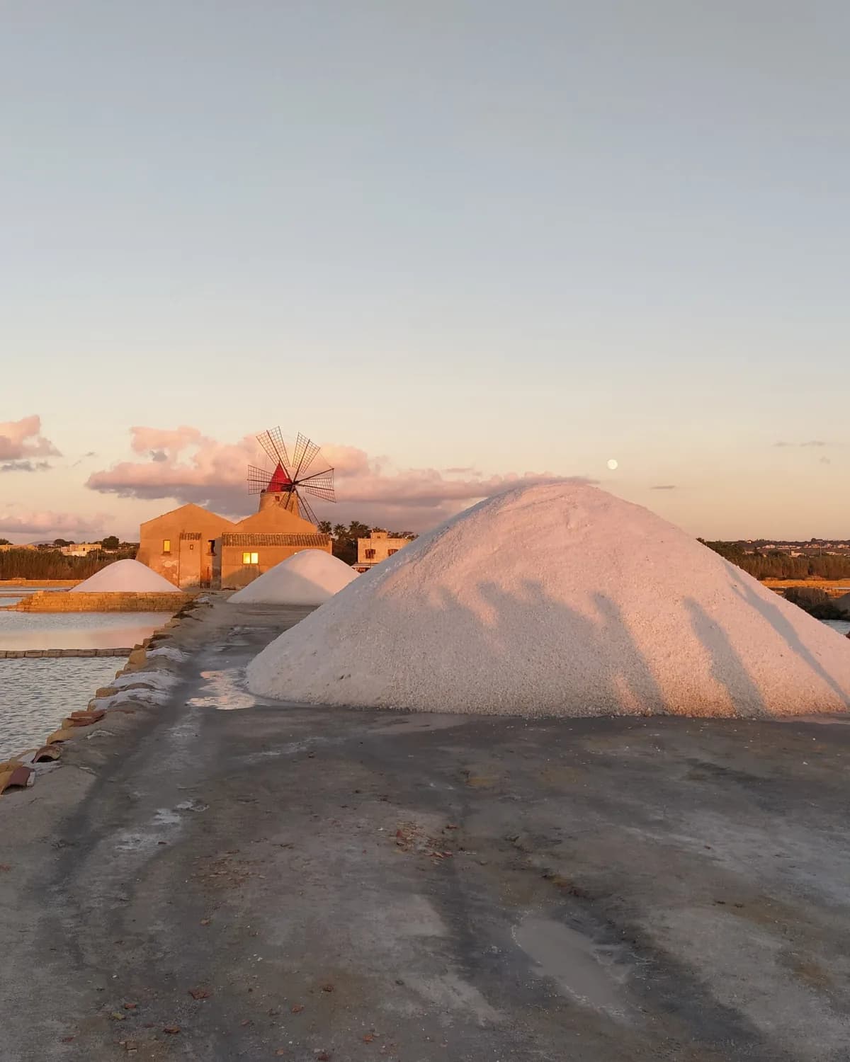 A large mound of salt beside a causeway at dusk, with a red-capped windmill and salt-works buildings behind it and the moon in a pale sky
