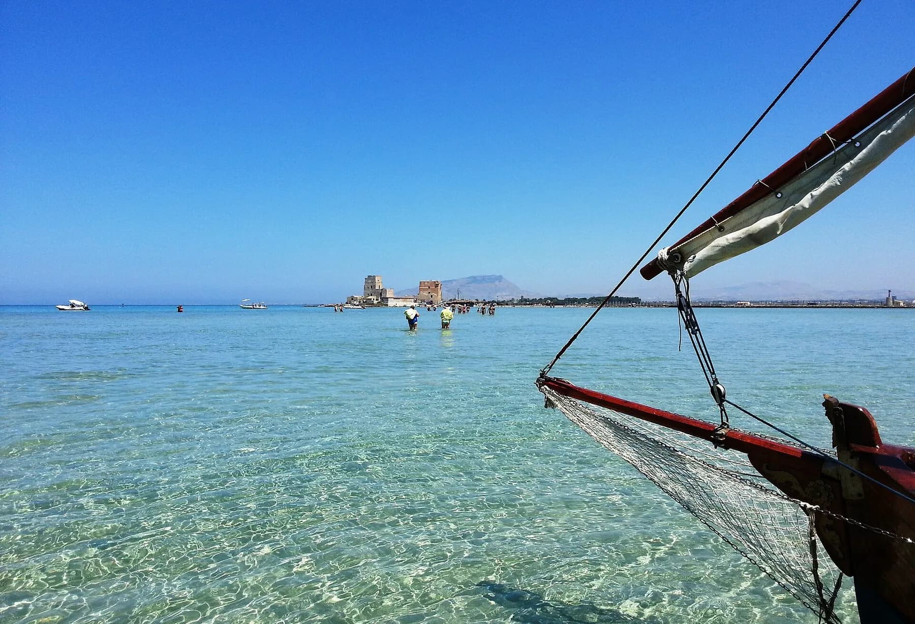 Clear shallow water of the lagoon with people wading towards a low island, a tower on the far shore and the bowsprit of a moored boat in the foreground