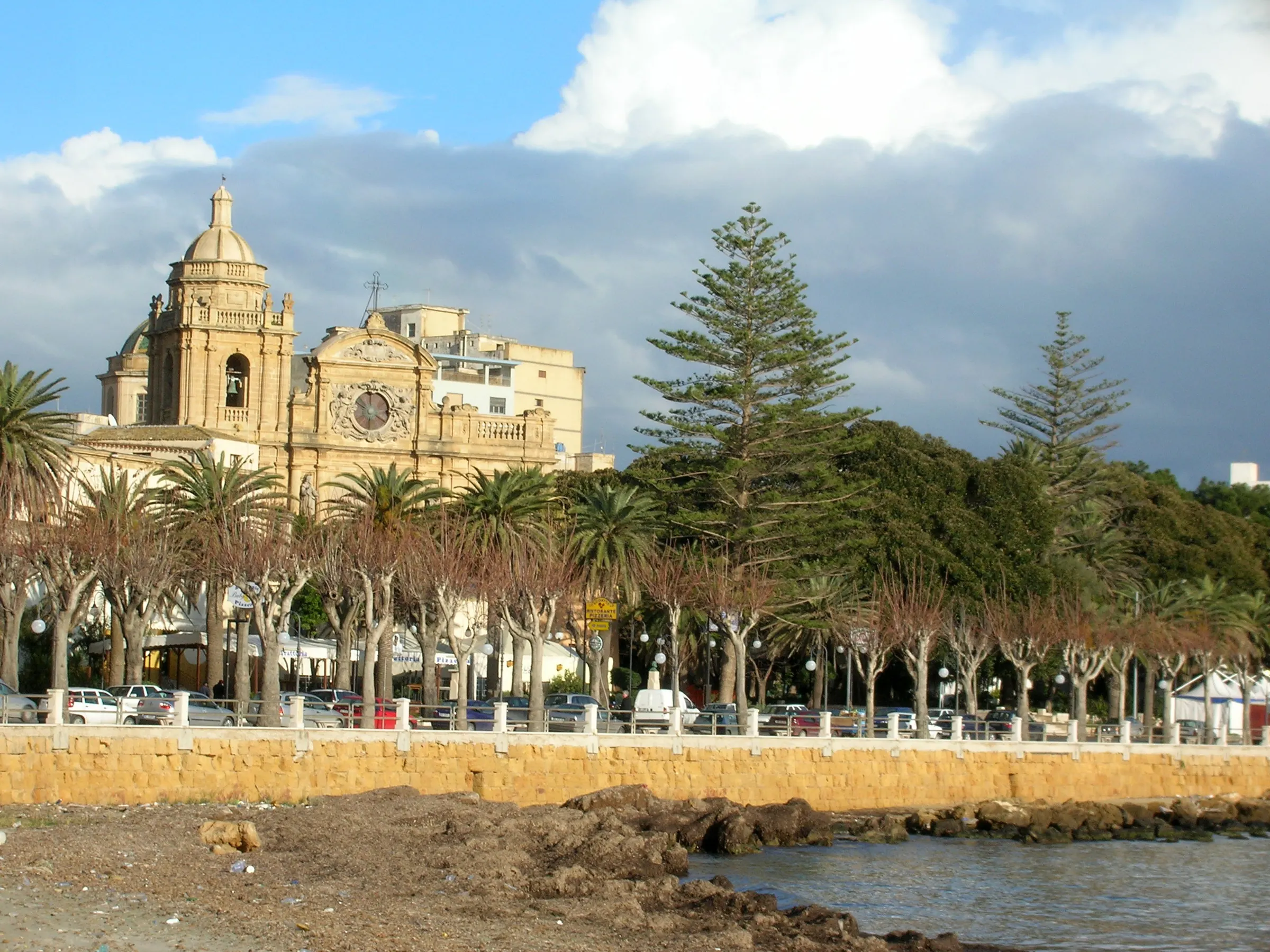 The Baroque façade and dome of the cathedral of Mazara del Vallo above a seafront lined with palms and pines