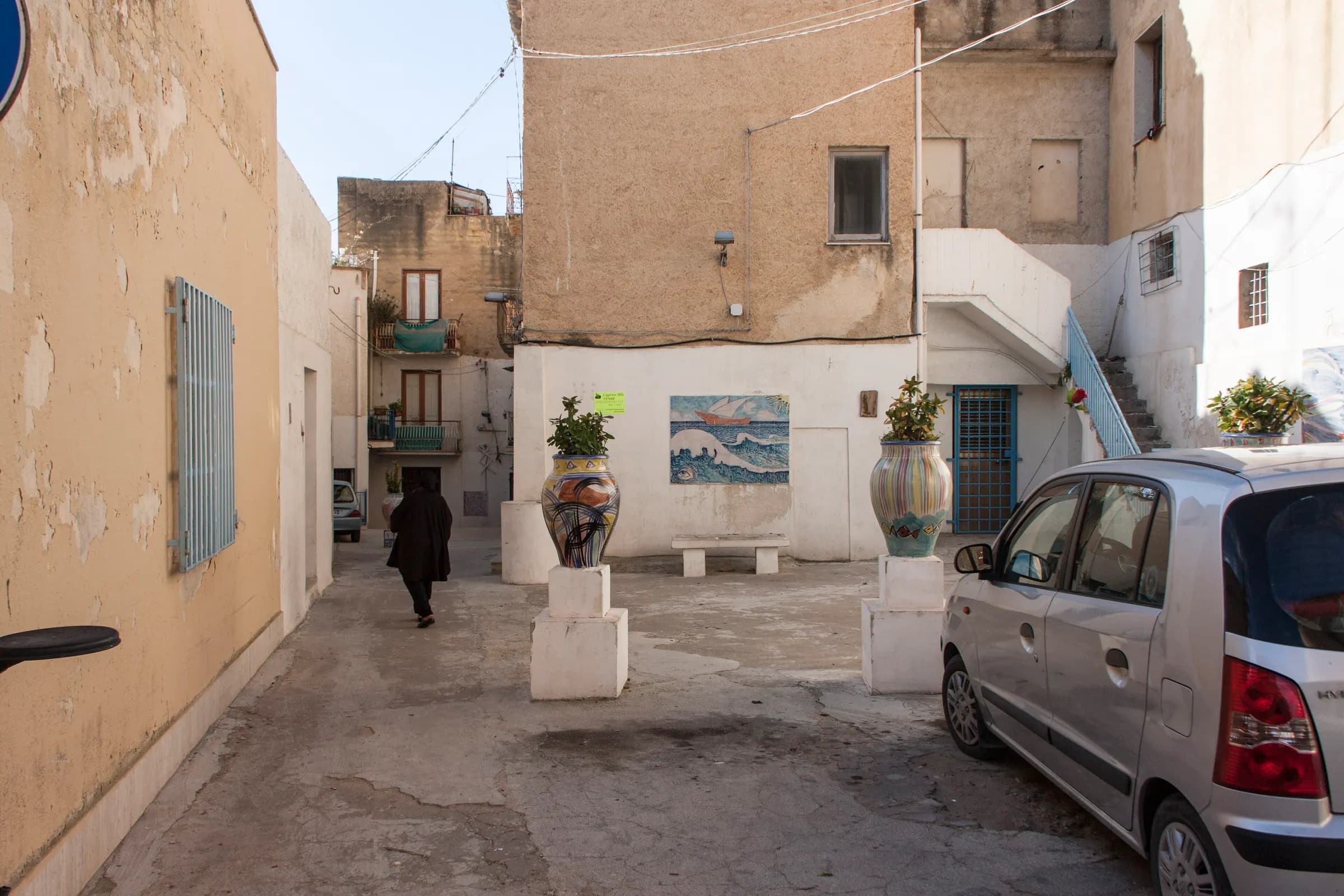 A lane in the Kasbah of Mazara del Vallo opening onto a small square with a tiled mural and painted ceramic pots