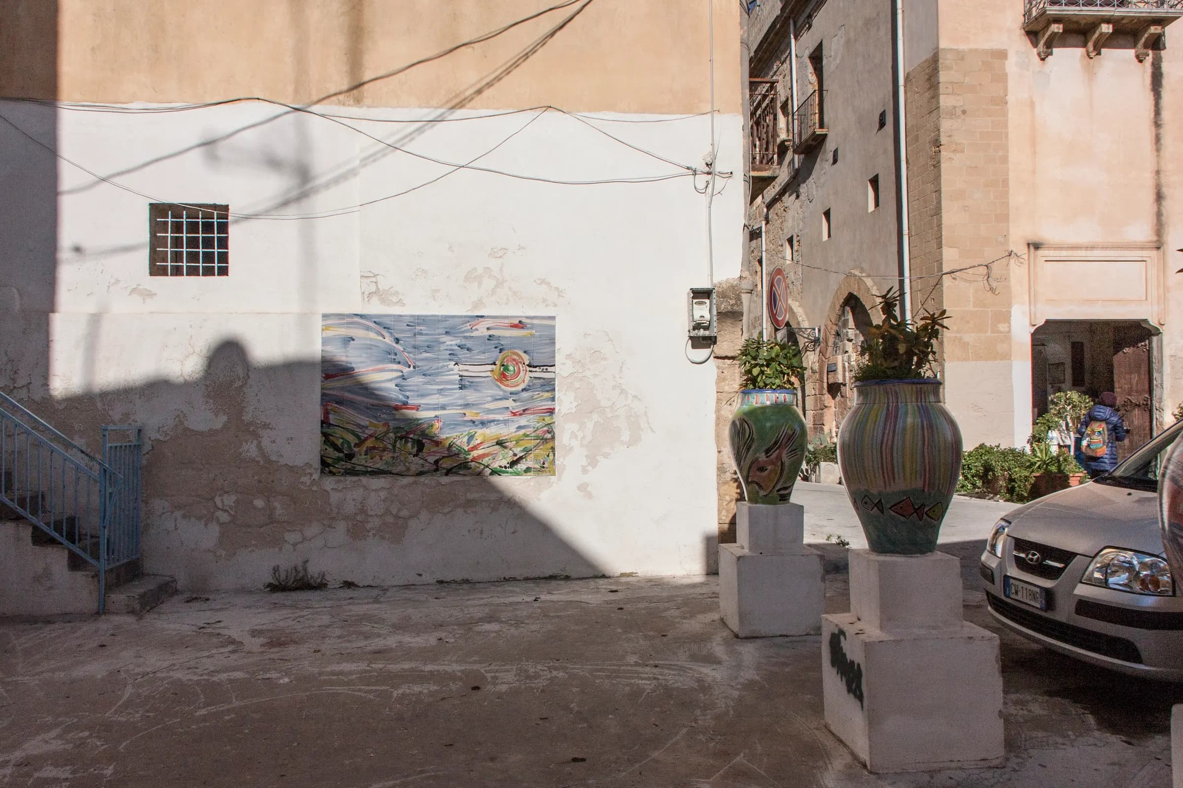 A small courtyard in the Kasbah of Mazara del Vallo, with a ceramic tile panel set into a whitewashed wall and painted pots on plinths