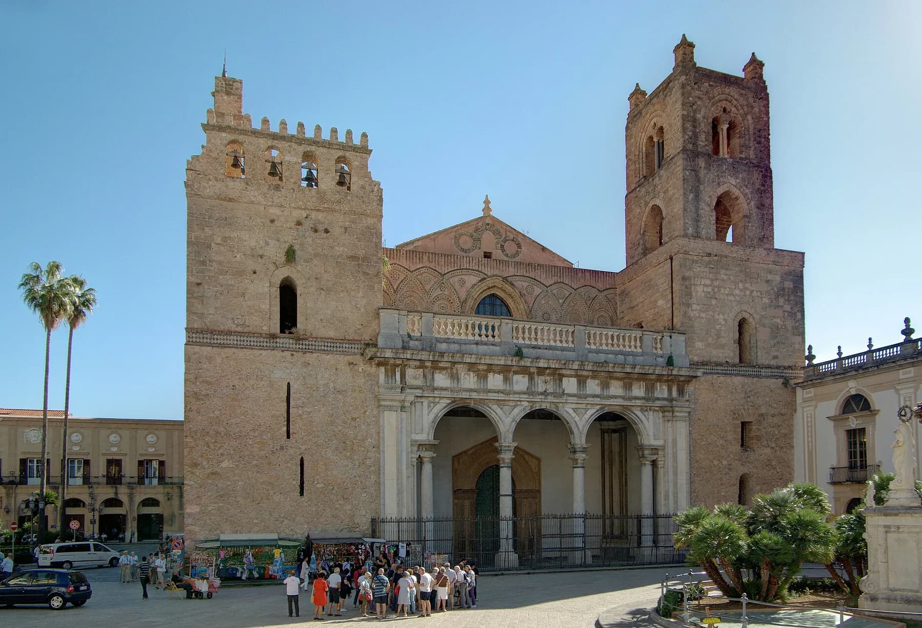 The façade of Monreale cathedral between its two square towers, with an arcaded porch and palm trees in the square