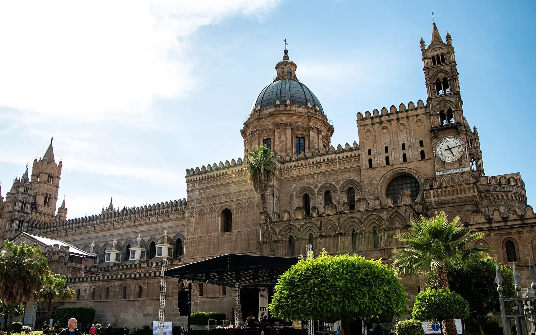 Palermo cathedral from the square, with its dome, crenellated walls, carved arcading and a clock tower, palms in the foreground