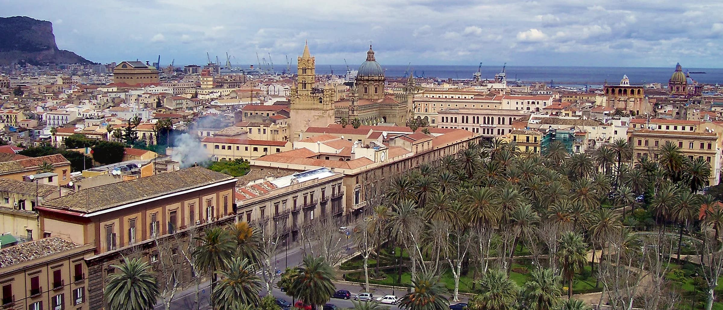 Palermo seen from above: tiled rooftops and palm trees spreading towards a cathedral tower and dome, with the port and the sea beyond