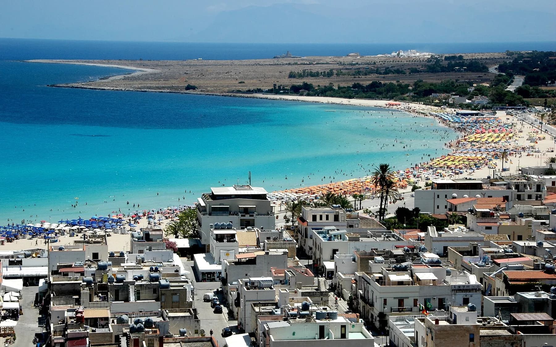 The town seen from above, white rooftops in the foreground and a crescent of beach lined with coloured umbrellas along a turquoise bay
