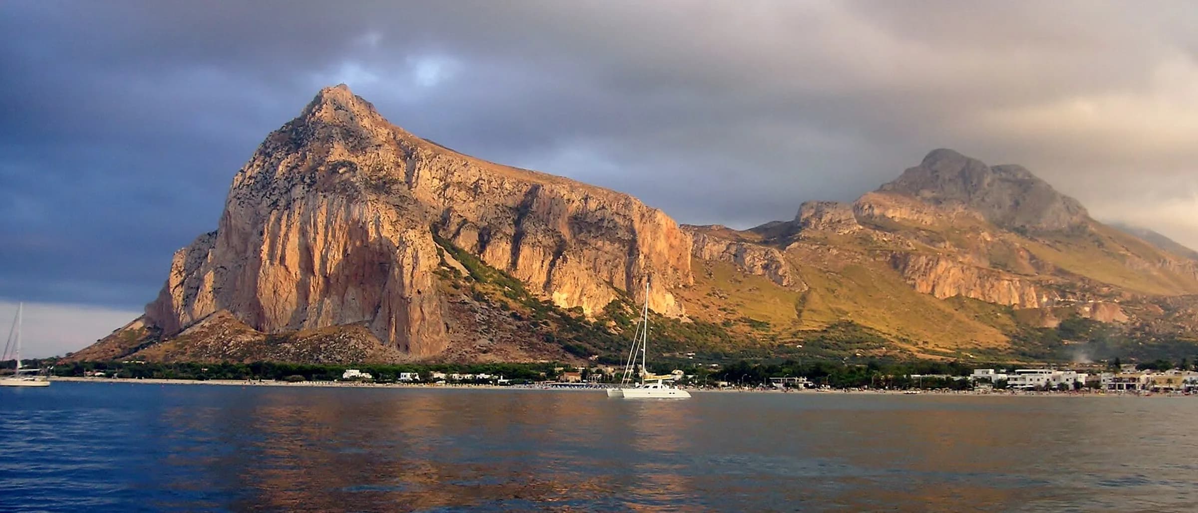 A steep rock face catching the late light above a low town along the shore, seen from the water with a catamaran moored offshore