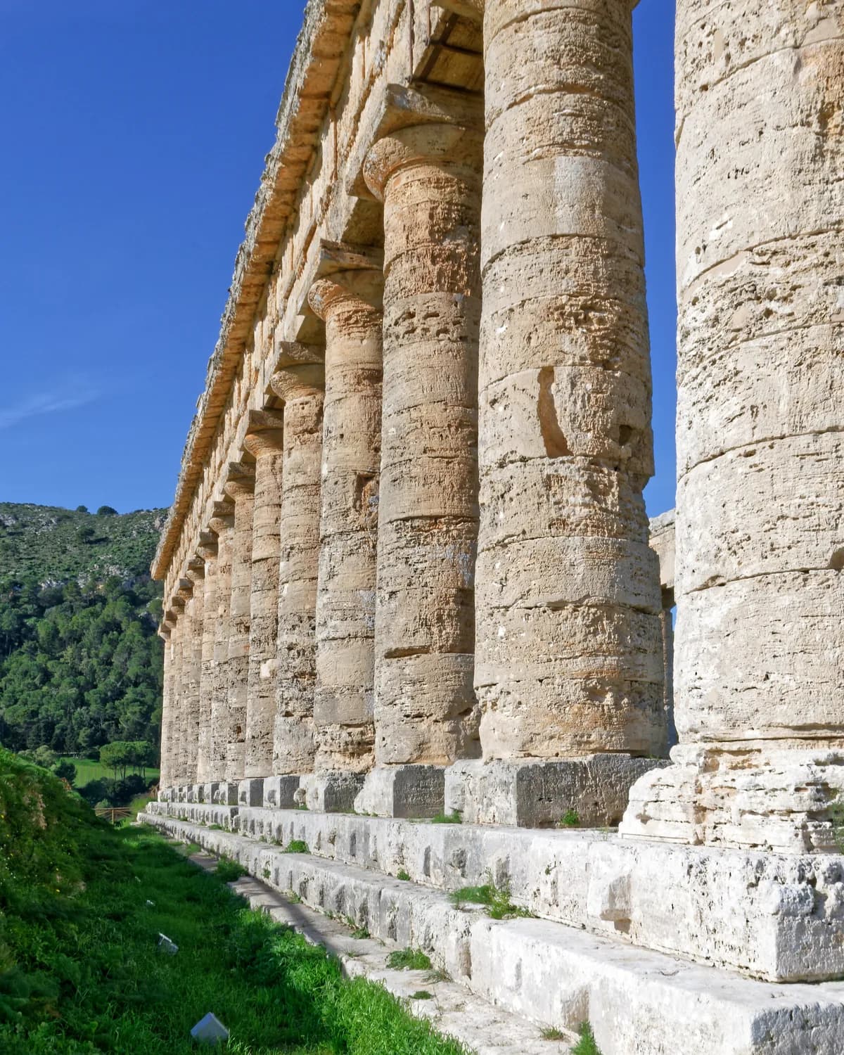 The temple colonnade seen close up from the side, weathered stone columns receding along the stepped base against a deep blue sky