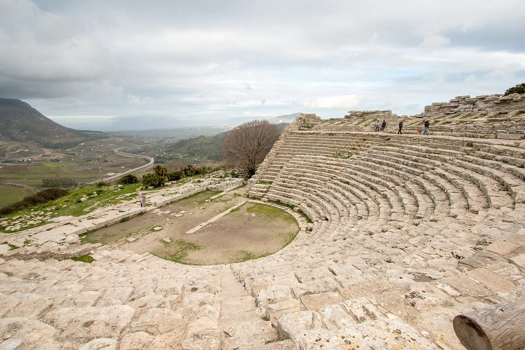The theatre seen from its upper rows, looking out past the tiers to a valley of fields and hills under a cloudy sky