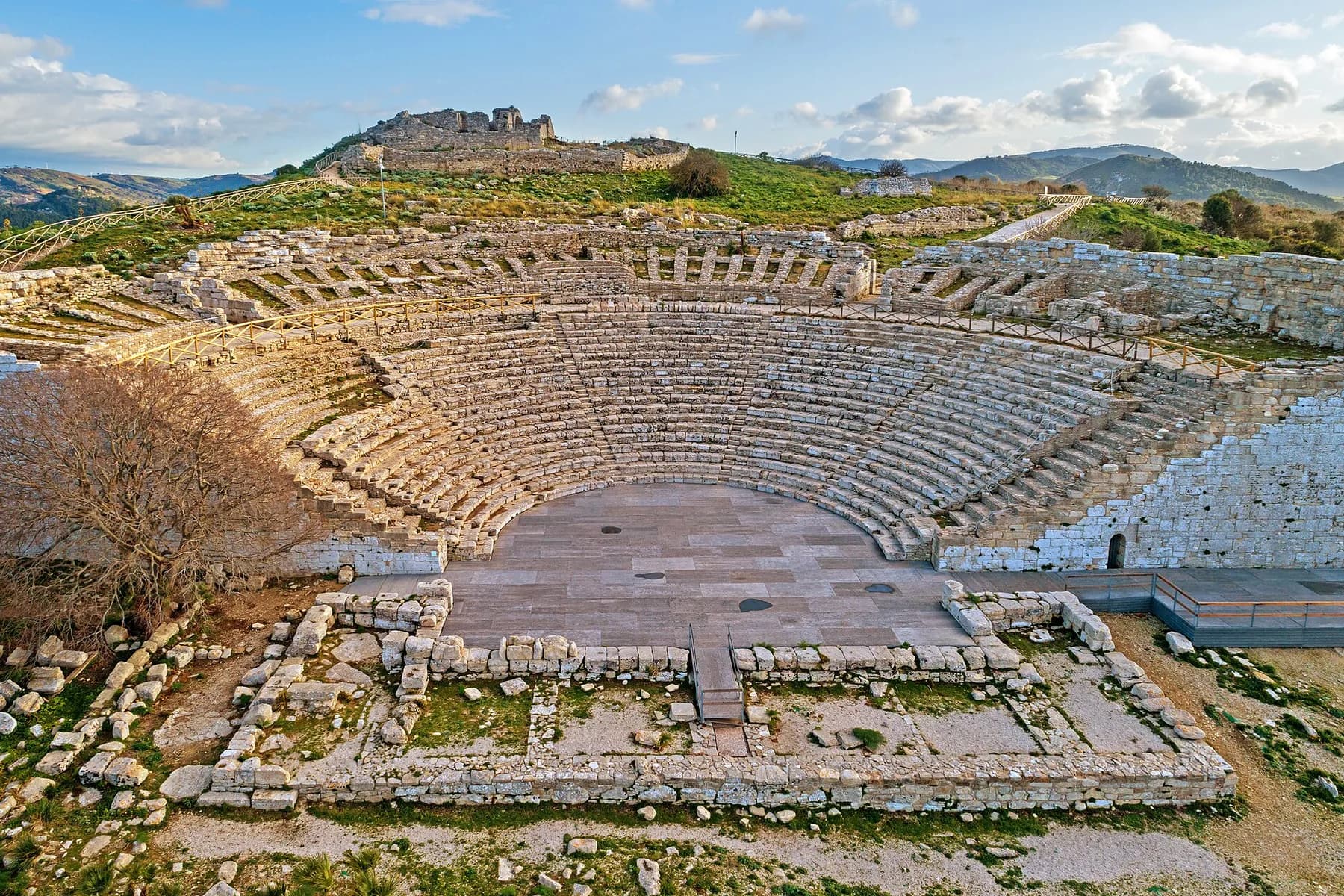 The ancient theatre at Segesta from above, its curved stone tiers wrapped around a paved stage, with ruins on the hilltop behind